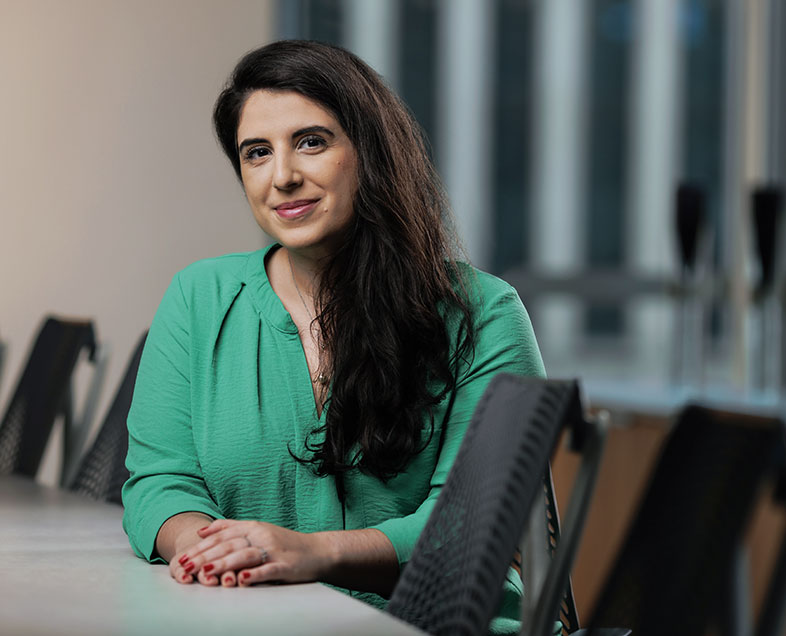 Marcella Costa Simões de Almeida  Mulher de cabelo castanho comprido. Veste uma blusa verde de mangas longas. Ela está sentada em uma cadeira, com as mãos entrelaçadas e apoiadas em uma mesa a sua frente, sorrindo.