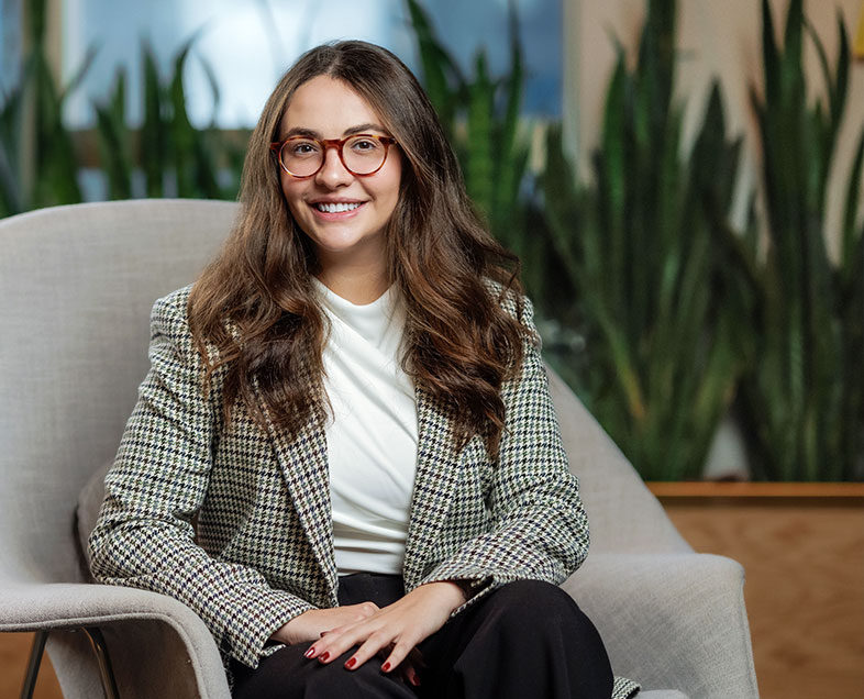 Marina Dalmaso Battistella  Mulher de cabelo castanho comprido e ondulado. Veste uma blusa branca, blazer branco com estampa quadriculada preta, calça preta e usa óculos. Ela está sentada em uma poltrona, com as pernas cruzadas e as mãos apoiadas sobre as pernas, olhando para frente e sorrindo.