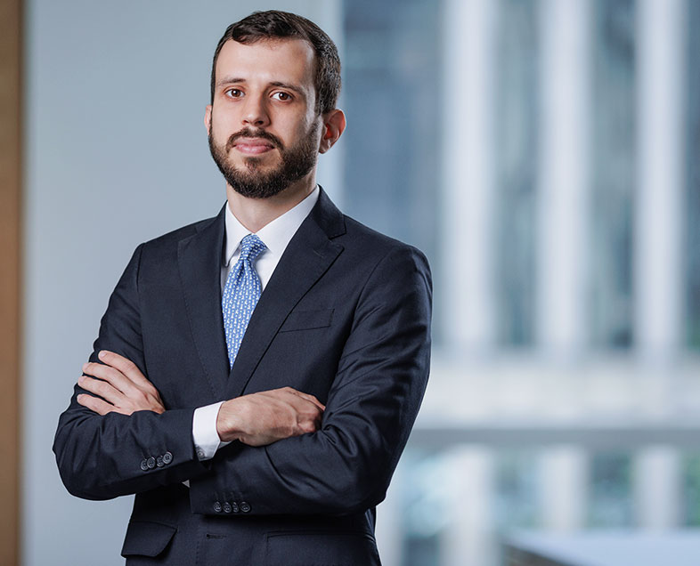 Matheus Holanda  Homem de cabelo preto curto, com barba e bigode curtos e da mesma cor. Veste uma camisa branca, terno preto e gravata azul clara com bolinhas brancas. Ele está em pé, de braços cruzados, olhando para frente.