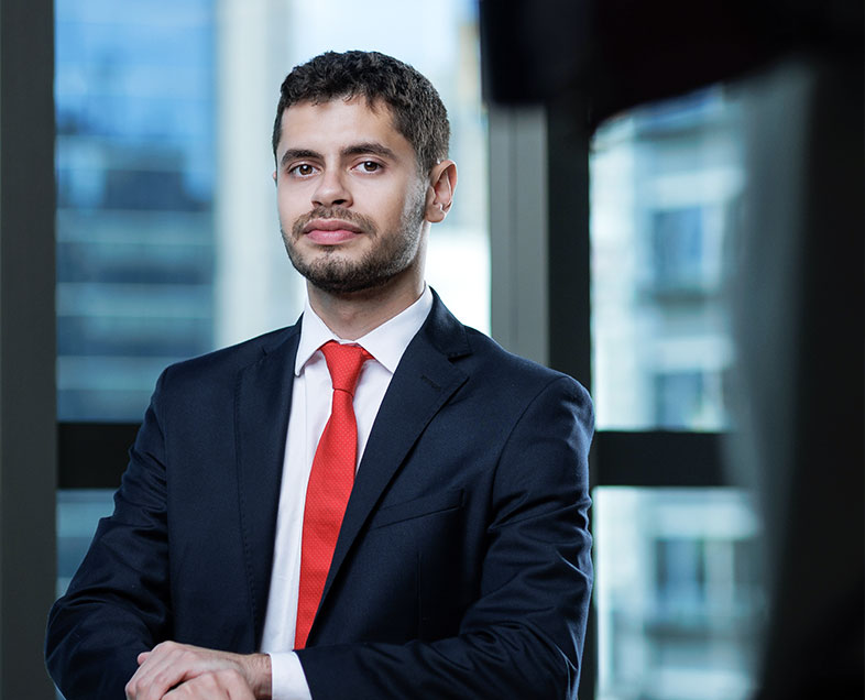 Murilo Guirao Souza  A man with short black hair and a short beard and moustache of the same color. He is wearing a white shirt, black suit and red tie. He is standing with his arms resting on a bench in front of him.