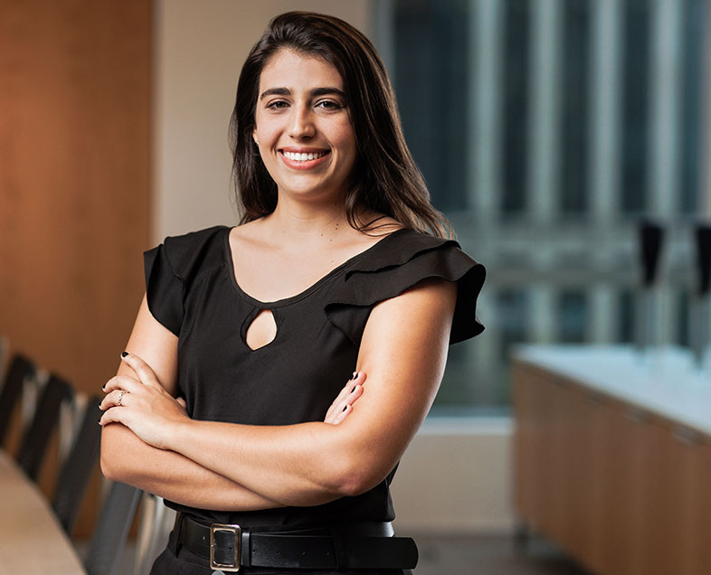 Nicole Gregolin  Mulher de cabelo preto comprido. Veste um vestido preto de mangas curtas com um cinto preto com fivela dourada. Ela está em pé de braços cruzados, olhando para frente e sorrindo.