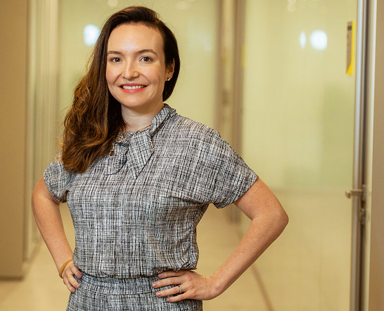 Manoela Vargas Nunes   Mulher de cabelo castanho na altura dos ombros. Veste um vestido de mangas curtas, cinza com estampa quadriculada, com um laço na gola. Ela esta em pé, com as mãos na cintura, sorrindo.