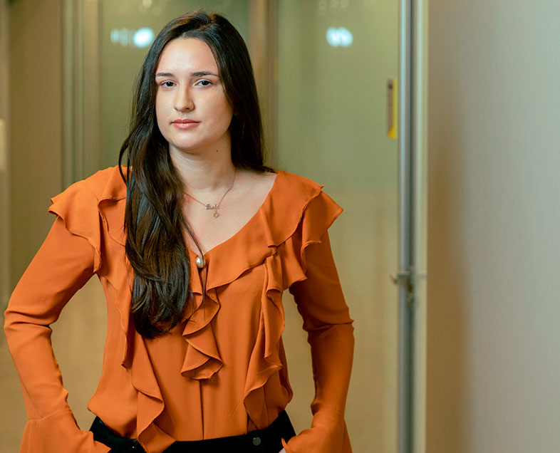 Paola Aranha Pacheco  Mulher de cabelo preto comprido. Veste uma blusa laranja de mangas compridas com babados na gola e na parte da frente da blusa e uma calça preta. Ela está em pé, com as mãos nos bolsos dianteiros da calça, olhando para frente.