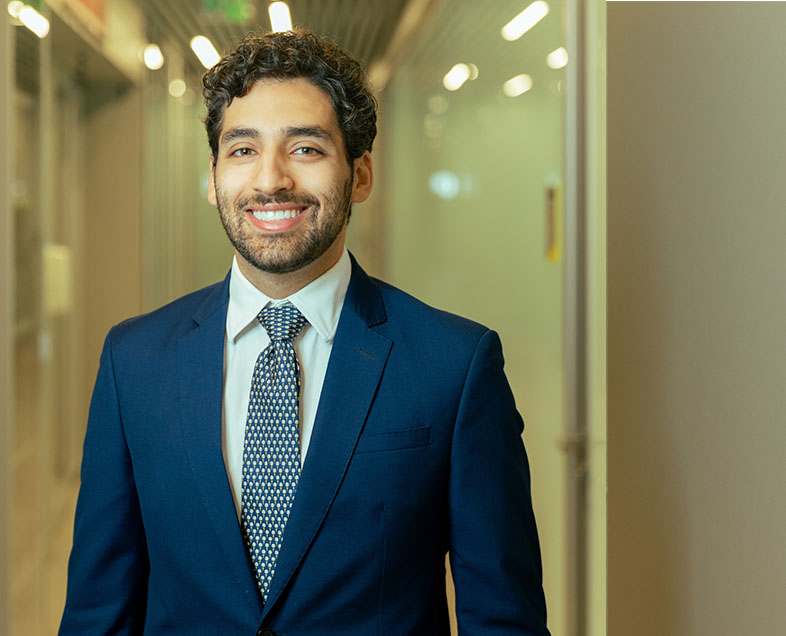 Pedro Gasparetto  A man with short curly brown hair, a short beard and mustache of the same color. He wears a white shirt, a blue suit and a blue tie with white polka dots. He is standing, looking straight ahead and smiling.