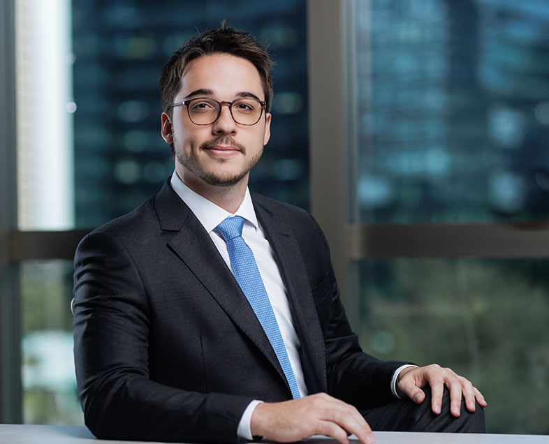 Pedro Perez Junqueira Sampaio Meirelles  Homem de cabelo preto curto com barba e bigode curtos da mesma cor. Veste uma camisa branca, terno preto, gravata azul clara e usa óculos. Ele está sentado em uma cadeira, virado para a direita, com as mãos apoiadas em uma mesa a sua frente, sorrindo.