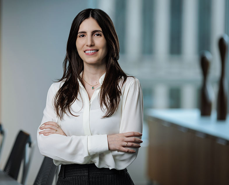 Paula Silveira Gonçalves  Mulher de cabelo preto comprido. Veste uma camisa branca de mangas longas e calça preta. Ela está em pé, de braços cruzados, olhando para frente e sorrindo.