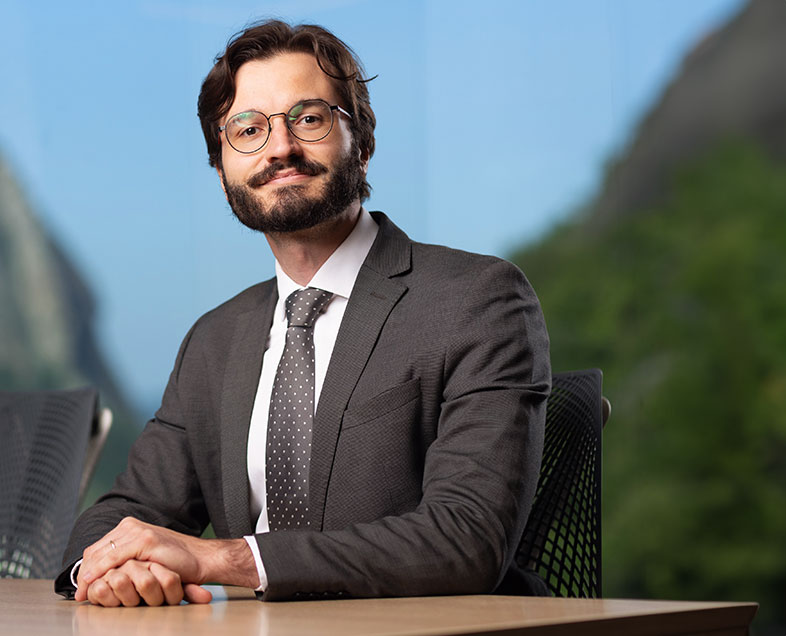 Raphael Barboza Correia  Homem de cabelo preto mediano e ondulado com barba e bigode curtos da mesma cor. Veste uma camisa branca, terno preto, gravata preta com bolinhas brancas e usa óculos. Ele está sentado em uma cadeira, virado para a esquerda, com as mãos apoiadas em uma mesa à sua frente frente, sorrindo.