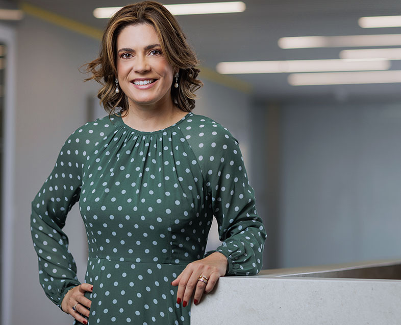 Roberta Danelon Leonhardt  Mulher de cabelo castanho na altura dos ombros. Veste um vestido verde de mangas compridas, com estampa de bolinhas brancas. Ela está em pé, apoiando seu braço esquerdo em uma bancada ao seu lado, olhando para frente e sorrindo. 