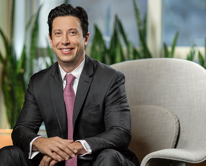 Raphael Zono  A man with short black hair. He is wearing a white shirt, a black suit and a burgundy tie. He is sitting in an armchair with his arms resting on his knees and his hands intertwined, looking straight ahead and smiling.  