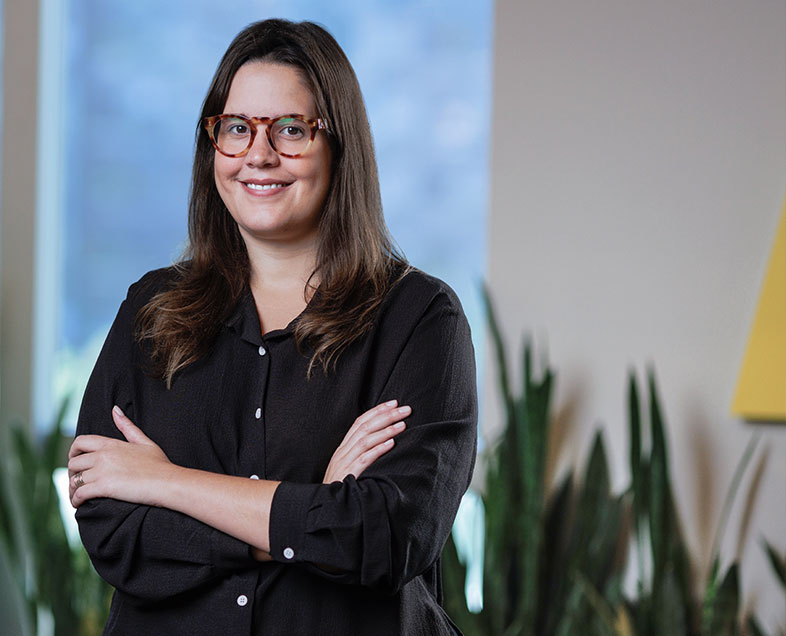 Raissa de Paula Xavier  Mulher de cabelo preto comprido. Veste uma camisa preta de meia manga e usa óculos. Ela está em pé, de braços cruzados, sorrindo.