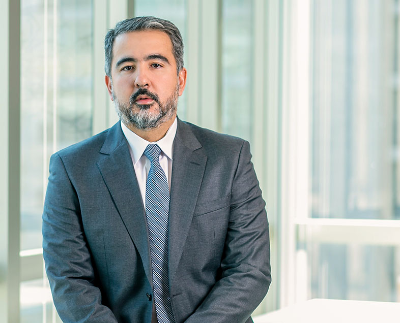 Rodrigo Seizo Takano  Homem de cabelo preto curto, meio grisalho, com barba e bigode curtos da mesma cor. Veste uma camisa branca, terno cinza e gravata azul. Ele está em pé, olhando para frente.