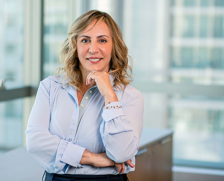 Andrea Giamondo Massei  Mulher de cabelo curto loiro. Veste uma camisa azul clara e uma calça azul escura. Ela está em pé, com a mão direita cruzada sob a barriga e a mão esquerda apoiada no queixo, está sorrindo e olhando para frente.