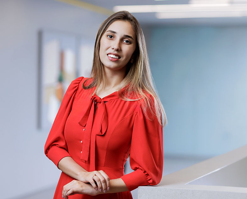 Sheyla Graziela Aparecida de Azevedo  Mulher de cabelo castanho comprido. Veste um vestido vermelho de meia manga bufante, com um laço na gola. Ela está em pé, com o braço esquerdo apoiado em uma bancada do lado direito, está olhando para frente e sorrindo.