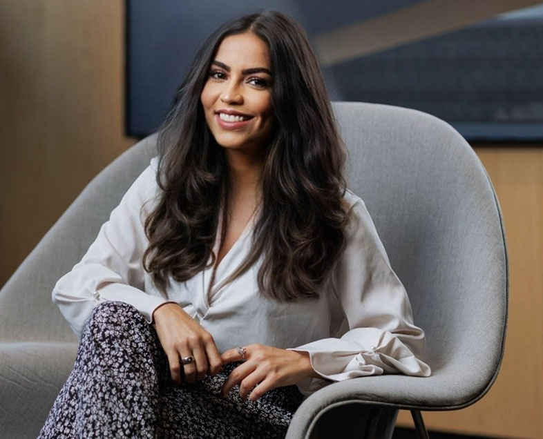 Stefanie Ólives  Mulher de cabelo preto comprido. Veste uma blusa branca de mangas longas e uma calça preta com estampa de flores brancas. Ela está sentada em uma poltrona, com as pernas cruzadas, o braço direito apoiado no joelho e o braço esquerdo apoiado no braço da poltrona, olhando para frente e sorrindo.