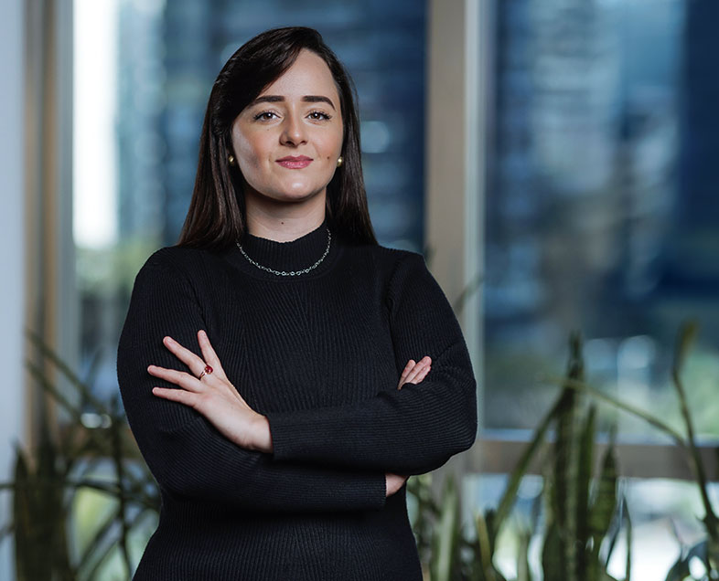 Tamiris Freire de Oliveira  Mulher de cabelo preto comprido. Veste um vestido preto de mangas longas e gola alta. Ela está em pé, de braços cruzados, olhando para frente.