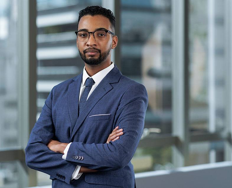 Vinícius Benevides Mota  Homem de cabelo preto curto com barba e bigode curtos da mesma cor. Veste uma camisa branca, terno azul escuro, gravata azul escura e usa óculos. Ele está em pé, de braços cruzados, olhando para frente.