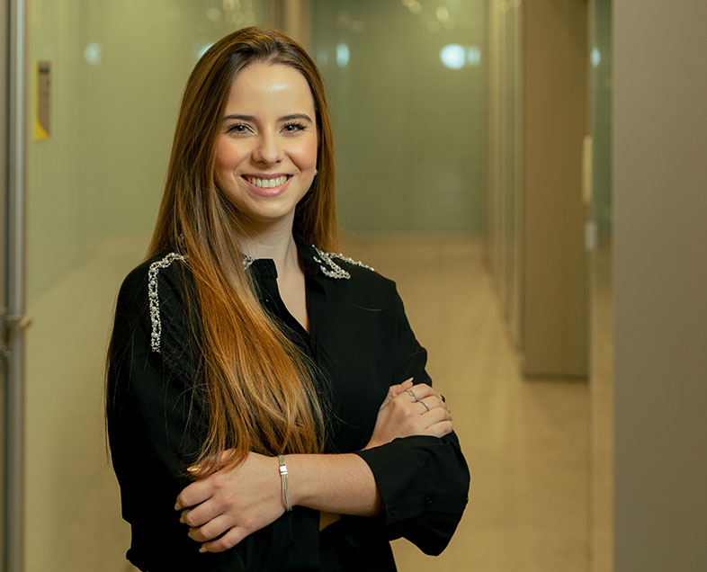 Victória Milaré Toledo Santos  Mulher de cabelo castanho comprido. Veste uma blusa preta de meia manga. Ela está em pé, virada para a direita, de braços cruzados, sorrindo e olhando para frente.