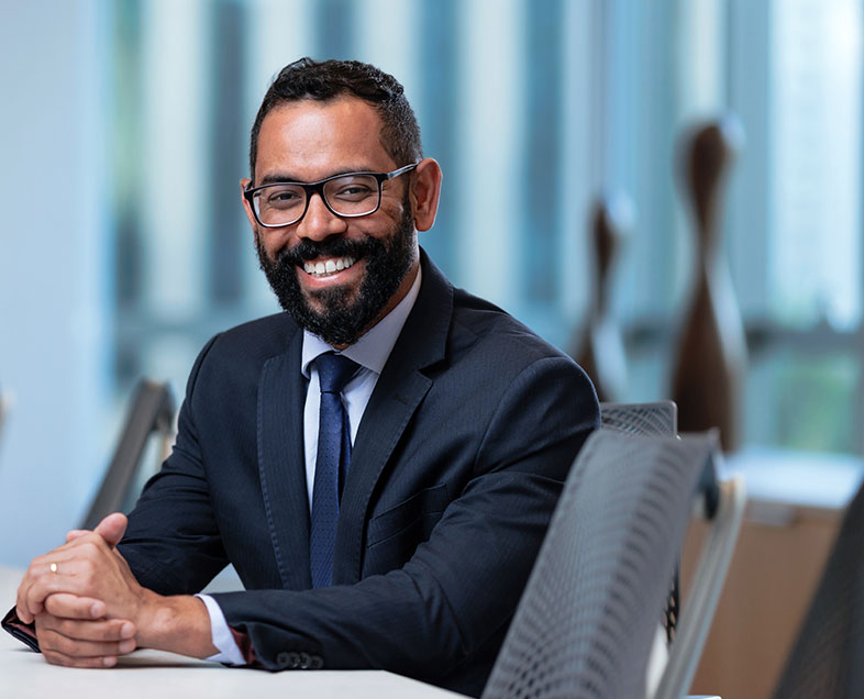 Wagner Mello dos Santos  Homem de cabelo preto curto com barba e bigode curtos da mesma cor. Veste uma camisa branca, terno preto, gravata azul escura e usa óculos. Ele está sentado em uma cadeira, com os braços apoiados em uma mesa a esquerda e as mãos entrelaçadas, olhando para frente e sorrindo.