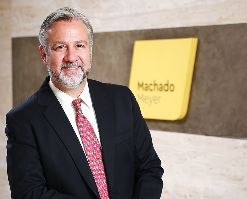 Leonardo Martins  A man with short gray hair, a short beard and mustache. He wears a white shirt, a black suit and a red tie with white polka dots. He is standing with his hands clasped in front of him, smiling.