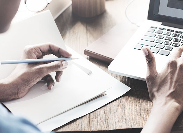 Pessoa digitando no notebook com a mão direita e escrevendo no papel com a mão esquerda