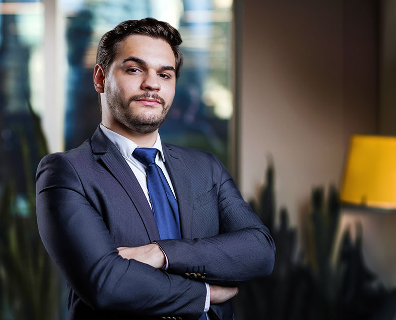 Caio Colognesi  Homem de cabelo curto castanho, com barba e bigode curtos e na mesma cor. Veste uma camisa branca, terno azul escuro e uma gravata azul marinho. Ele está em pé, virado para a direita, de braços cruzados e olhando para frente