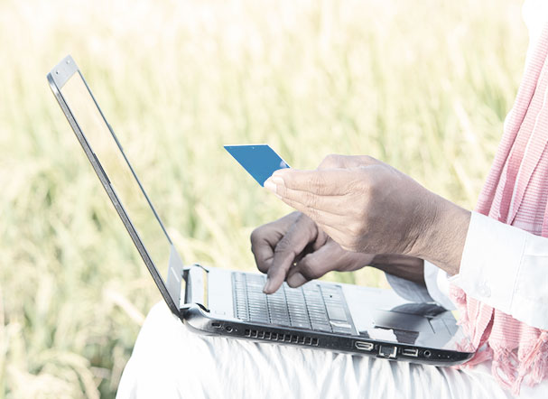 Agricultor sentado próximo de sua plantação, usando roupa branca, usando notebook para fazer pagamento online com cartão de crédito